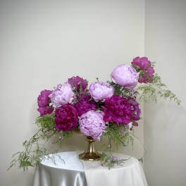 Low compote arrangement of pink and magenta peonies with trailing greenery in a gold pedestal vase.