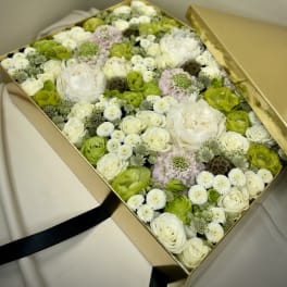 Square gold box filled with white, green, and pale lavender flowers with the lid set to the side