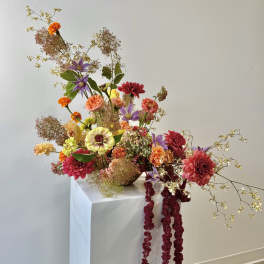 Tall asymmetrical arrangement of warm-toned dahlias and mixed flowers in a low glass bowl on a white pedestal.
