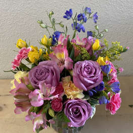 Mixed bouquet of purple roses, pink alstroemeria, and yellow blooms in a glass vase