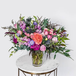 Lush arrangement of pink, purple, and peach flowers in a clear glass vase on a small round table.