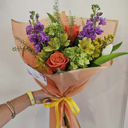 Handheld bouquet of purple, orange, yellow, and green flowers wrapped in peach paper