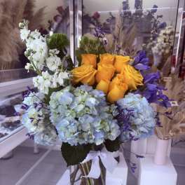 Vase arrangement of yellow roses, blue hydrangeas, white blooms and purple accents in a clear glass cylinder