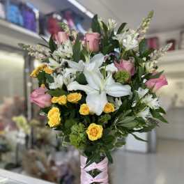 Tall arrangement of white lilies, pink roses, and yellow spray roses in a glass vase wrapped with pink ribbon