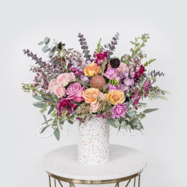 Mixed bouquet of pink, peach, and purple flowers in a speckled white vase