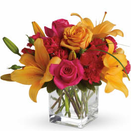 Bright arrangement of orange lilies, pink roses, and red carnations in a clear glass cube vase