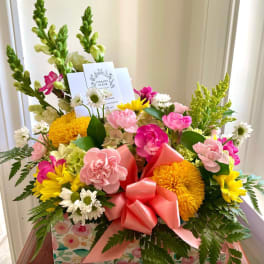 Colorful box arrangement of pink carnations, yellow mums, and white daisies with a pink bow and card.
