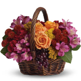 Low basket arrangement of orange roses, pink alstroemeria, red blooms, and yellow flowers with grapes.
