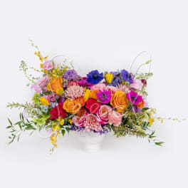 Low centerpiece of bright pink, yellow, orange, and blue flowers in a white vase