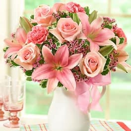 Pink lilies, roses, and carnations arranged in a white pitcher with a pink ribbon bow.