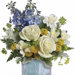 Short arrangement of white roses, yellow mums, and blue delphinium in a blue baby block vase