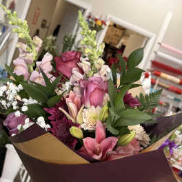 Hand-tied bouquet of pink roses, lilies, and mixed blooms wrapped in burgundy paper with a gold ribbon.