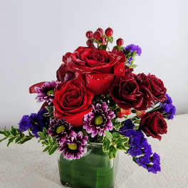 Red roses and purple daisies arranged in a square glass vase