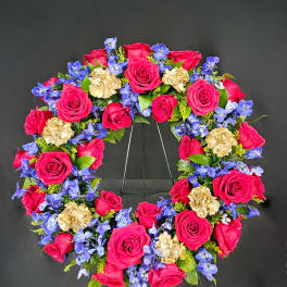 Heart-shaped wreath of pink roses, blue flowers, and gold carnations on a stand
