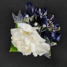 White carnation corsage with navy ribbon loops, tiny purple accents, and jeweled picks on a dark background.