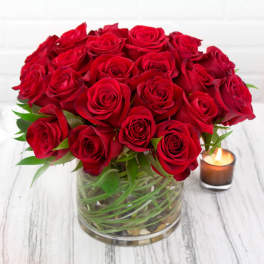 Low round arrangement of red roses in a clear glass vase beside a lit candle