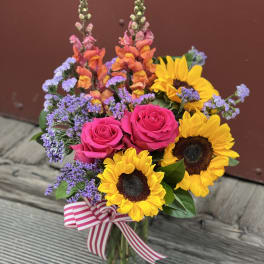 Bouquet of pink roses, sunflowers, and purple filler in a glass vase with a striped ribbon.