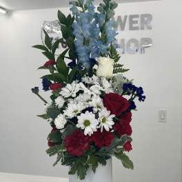 Tall floral arrangement with blue delphinium, white daisies, red roses, and carnations in a white vase
