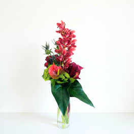 Tall modern arrangement of red orchids, calla lilies, and anthurium with large glossy green leaves in a clear glass vase
