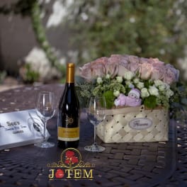 Pink rose arrangement in a woven basket with a small plush toy and wine bottle nearby