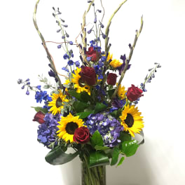 Tall bouquet of sunflowers, red roses, and blue flowers in a glass vase