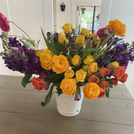 Colorful mixed bouquet in a white bucket vase with yellow, orange, pink, and purple blooms.
