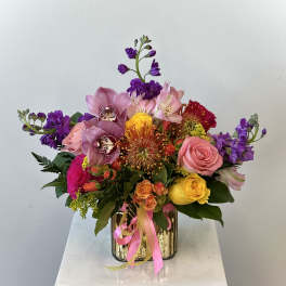 Colorful bouquet of roses, orchids, and mixed blooms in a gold vase with pink ribbon