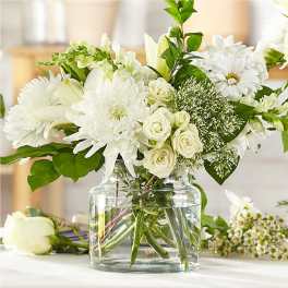 White roses, lilies, daisies, and mums arranged in a clear glass vase with water on a light surface
