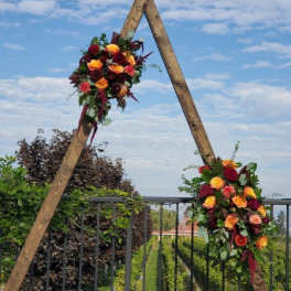 Autumn-toned floral sprays of red and yellow roses on a rustic wooden triangle frame outdoors.