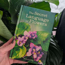Hand holding a book with purple flowers on the cover in front of large green leaves