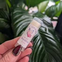 Hand holding a lip balm tube in front of large green leaves.
