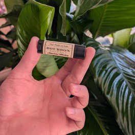Hand holding a bourbon lip balm in front of large green leaves.
