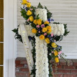 White floral cross on an easel with yellow roses and a memorial ribbon