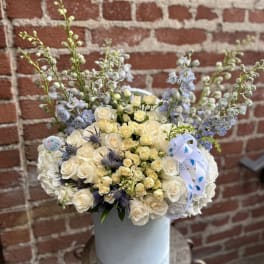 Tall hatbox arrangement of ivory roses and blue flowers with baby boy ribbon accents
