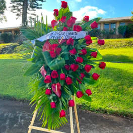 Standing spray of red roses and tropical blooms on an easel with a memorial banner.