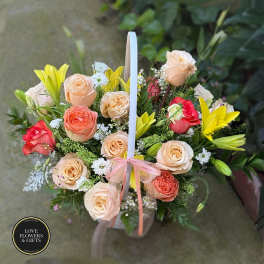 Basket arrangement of peach roses, red roses, and yellow lilies with a ribbon bow on the handle