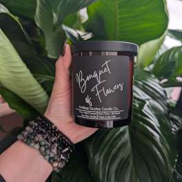 Hand holding a black candle jar labeled "Bouquet of Flowers" in front of large green leaves.