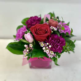 Compact arrangement of red rose, purple carnations and pink blooms in a pink cube vase with a glitter heart pick