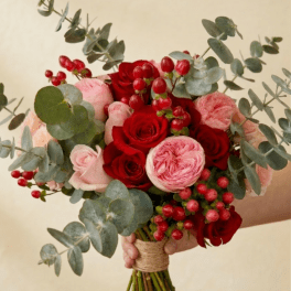 Hand-tied bouquet of red and pink roses with red berries and eucalyptus, held against a beige background.