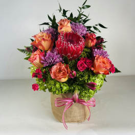 Mixed bouquet with roses, purple mums, and a red protea in a burlap-wrapped container