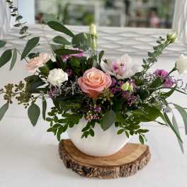Low pink and white flower arrangement with roses, orchids, and lisianthus in a white bowl on a wood slice base