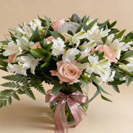 Round vase arrangement of peach roses and white alstroemeria with a pink ribbon bow.