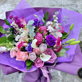 Hand-tied bouquet of pink and purple flowers with white calla lilies wrapped in purple paper.