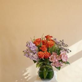 Orange roses with pink and lavender flowers in a clear glass vase on a light background