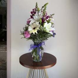 Tall mixed bouquet of white lilies and daisies with purple and pink flowers in a clear vase with a purple bow.
