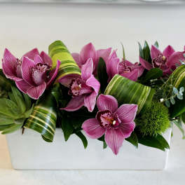 Low arrangement of pink orchids with succulents and rolled striped leaves in a white rectangular container