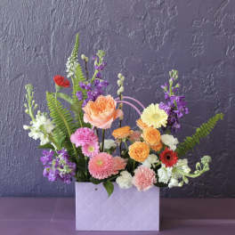 Colorful mixed flower arrangement in a lavender gift bag-style container against a purple wall