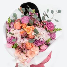 Large bouquet of pink, peach, and lavender flowers wrapped in white paper, held by a person in pink.