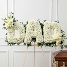White floral tribute spelling DAD on an easel beside a casket