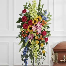 Tall standing floral spray with red roses, sunflowers, lilies, and blue accents beside a casket